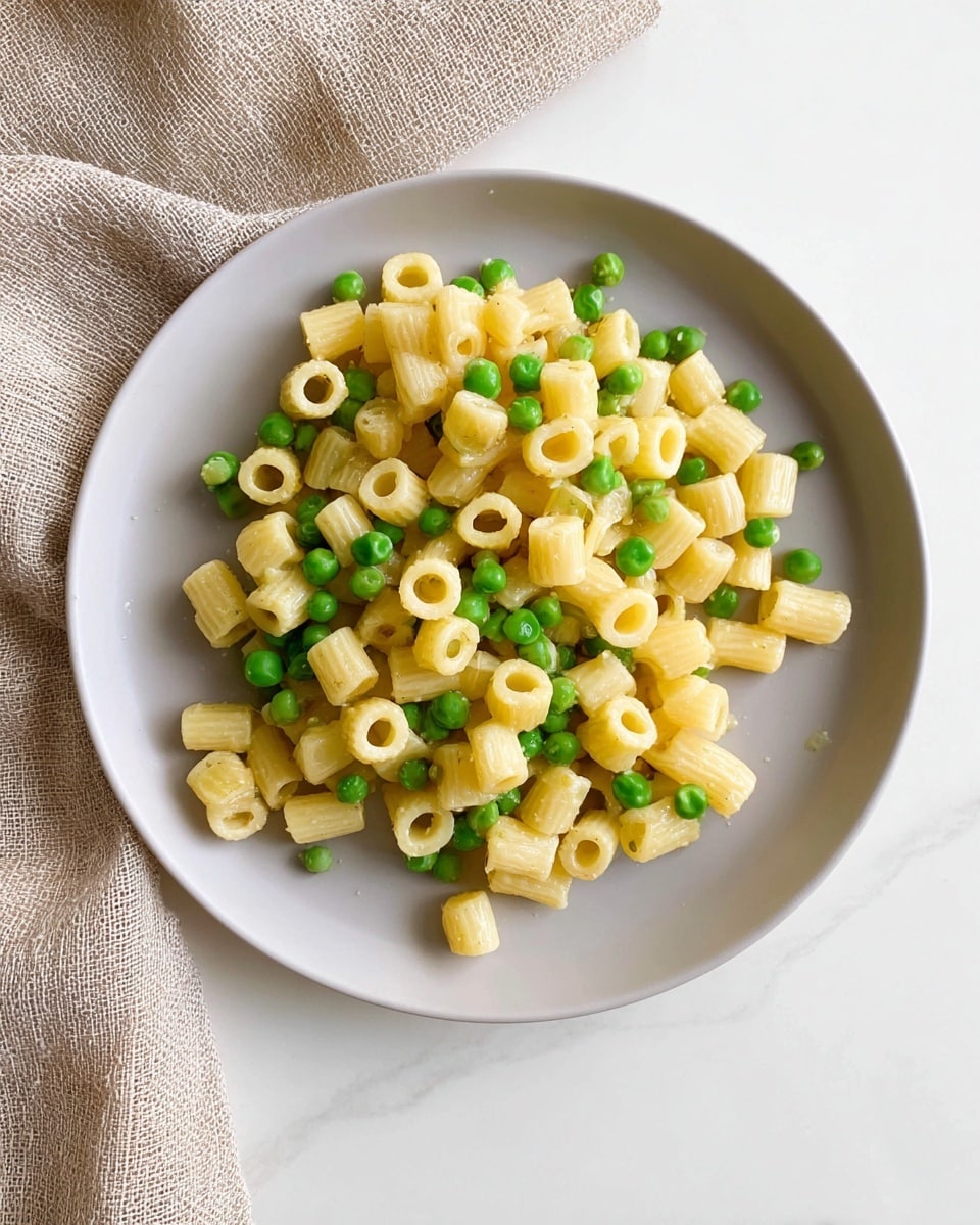 A close-up view of a single layer of short tubular pasta mixed evenly with bright green peas, all lightly coated in a creamy sauce that gives a slight shine to the pale yellow pasta. The pasta and peas sit centered on a round white plate with a smooth matte finish. The plate is placed on a white marbled texture, with a beige woven cloth softly draped in the background near the upper edge. Photo taken with an iphone --ar 4:5 --v 7