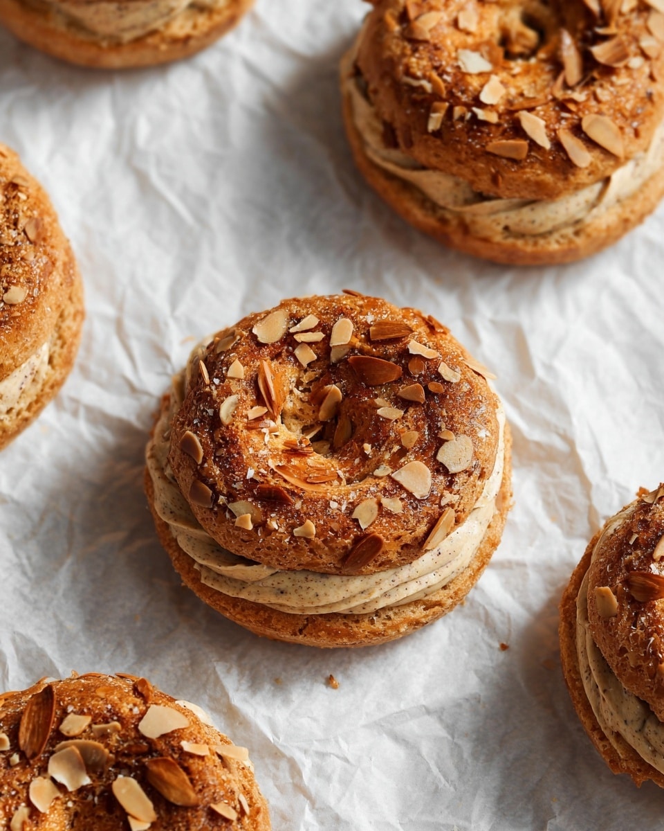 The image shows several round pastries with a rough, golden-brown top layer sprinkled with toasted almond slices. Each pastry is made of two main layers: the top layer is a baked, textured dough with a shiny brown finish, and below it is a thick swirl of light beige cream with specks, visible through the gap in the middle. The pastries rest directly on a crinkled white parchment paper with a soft, neutral look. The whole scene has a soft, natural light that highlights the textures and warm tones of the pastries. photo taken with an iphone --ar 4:5 --v 7