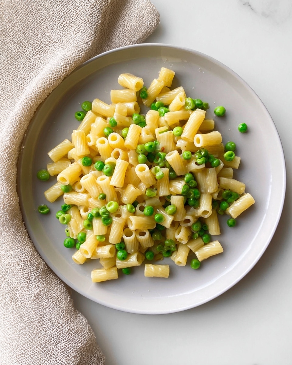 A close-up view of a simple pasta dish on a white plate, featuring small tube-shaped pasta mixed evenly with bright green peas. The pasta pieces are pale yellow with a slightly glossy texture, scattered naturally across the plate, with some peas peeking through and adding a pop of green color. The plate is placed on a white marbled surface with a beige textured cloth softly folded near the top edge, creating a clean and minimal look. Photo taken with an iphone --ar 4:5 --v 7