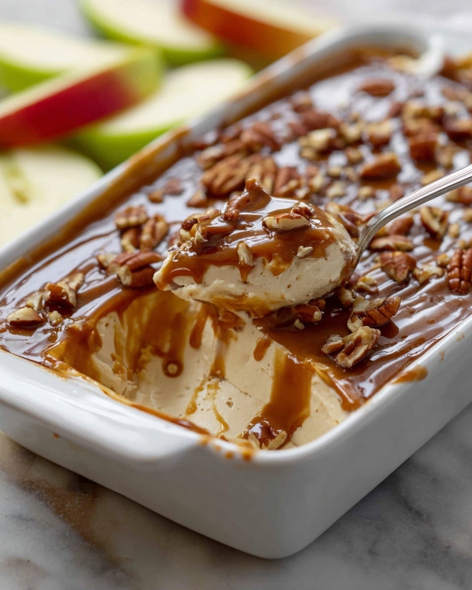 A white rectangular dish holds a creamy three-layer dessert. The bottom layer is light beige and smooth, the middle layer is thicker and creamy white, and the top layer is a glossy dark caramel sauce sprinkled with chopped pecans. A spoon scoops through the layers, mixing the creamy and caramel parts. The dish is on a white marbled surface with green and red apple slices nearby. Photo taken with an iphone --ar 4:5 --v 7