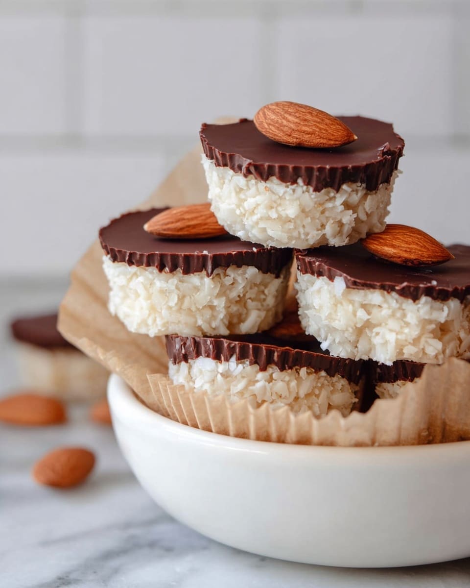 The image shows a stack of small round treats in a white bowl lined with light brown paper cups, placed on a white marbled surface. Each treat has two layers: a thick, textured bottom layer made of white shredded coconut, and a smooth, dark brown chocolate layer on top. On top of the chocolate layer, there is a single whole almond placed in the center of each treat. Some treats are stacked neatly, while others are leaning, showing the different layers clearly. The whole scene has a soft and clean look with a blurred white tile background. photo taken with an iphone --ar 4:5 --v 7