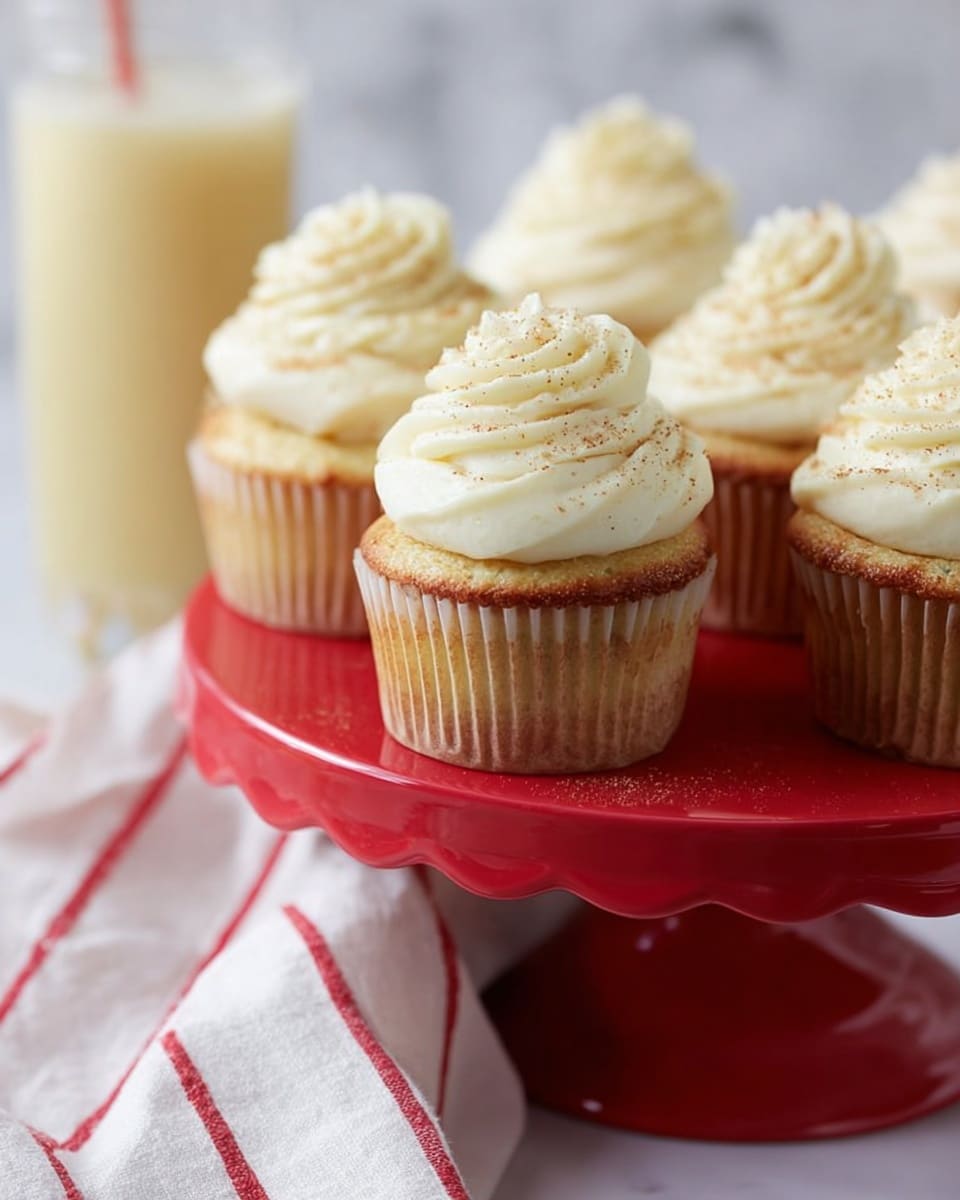 The image shows several vanilla cupcakes with light golden-brown bases, each topped with a thick, tall swirl of creamy white frosting that has fine specks of vanilla or spice sprinkled on top. The cupcakes are placed on a shiny red cake stand with scalloped edges, and the background includes a glass filled with a pale yellow drink. A white cloth with red stripes is seen draped casually under the cake stand, all set against a white marbled texture. photo taken with an iphone --ar 4:5 --v 7