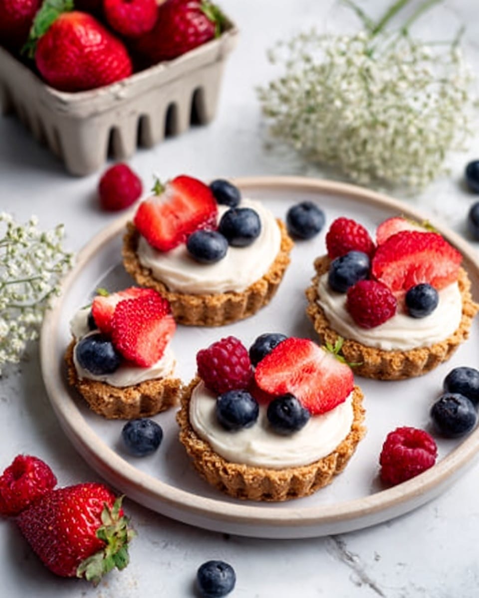 The image shows four small tartlets on a round white plate placed on a white marbled surface. Each tartlet has a golden brown crust base with a smooth, creamy white filling on top. They are decorated with fresh fruits: bright red strawberries, small dark blueberries, and a few red raspberries, arranged neatly with one or two strawberry slices and a couple of blueberries on each tart. Next to the plate, on the white marbled surface, there are extra fresh strawberries and blueberries scattered for decoration, along with a bunch of small white flowers and a white container filled with strawberries in the background. Photo taken with an iphone --ar 4:5 --v 7