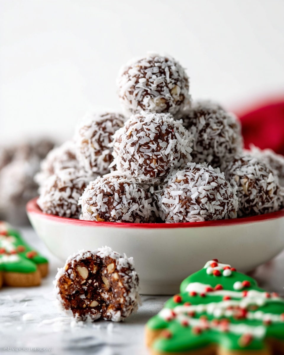 A pile of round chocolate balls covered with white shredded coconut is stacked in a pyramid shape inside a white bowl with a red outer edge, showing a rough textured surface with chunks of nuts visible through the coconut flakes. One chocolate ball lies in front of the bowl on a white marbled surface. Near it are blurry green Christmas tree-shaped cookies decorated with white and red icing and small colorful sprinkles. The background is bright white, making the colors and textures of the treats stand out. photo taken with an iphone --ar 4:5 --v 7