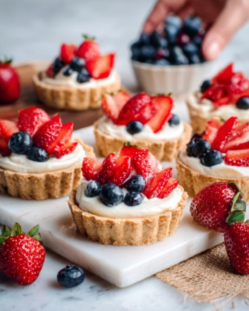 The image shows small tarts with three visible layers in each. The bottom layer is a crisp golden brown tart crust with a textured edge. The middle layer is smooth, creamy white filling that fills the crust almost to the top. The top layer is made of fresh strawberries and blueberries, cut and whole, arranged neatly on the cream, adding red and dark blue colors with a shiny, juicy appearance. The tarts are placed on a white tray on a white marbled surface, with some whole strawberries and blueberries scattered around for decoration. A woman's hand is gently holding one tart on the right side. Photo taken with an iphone --ar 4:5 --v 7