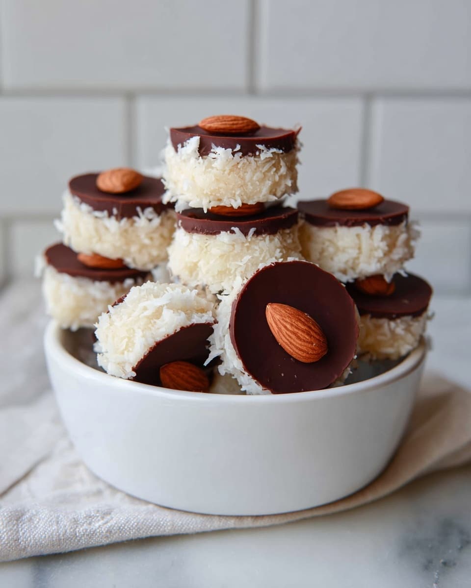 A white bowl is filled with a stack of small round treats that have two layers: a bottom thick layer of white shredded coconut texture and a top smooth dark brown chocolate layer. Each treat is topped with a single almond placed in the center, adding a contrasting brown color. The treats are arranged unevenly, some standing upright and others tilted, creating a natural, rustic pile. The background shows a white tiled wall, and the bowl is placed on a white marbled surface. photo taken with an iphone --ar 4:5 --v 7