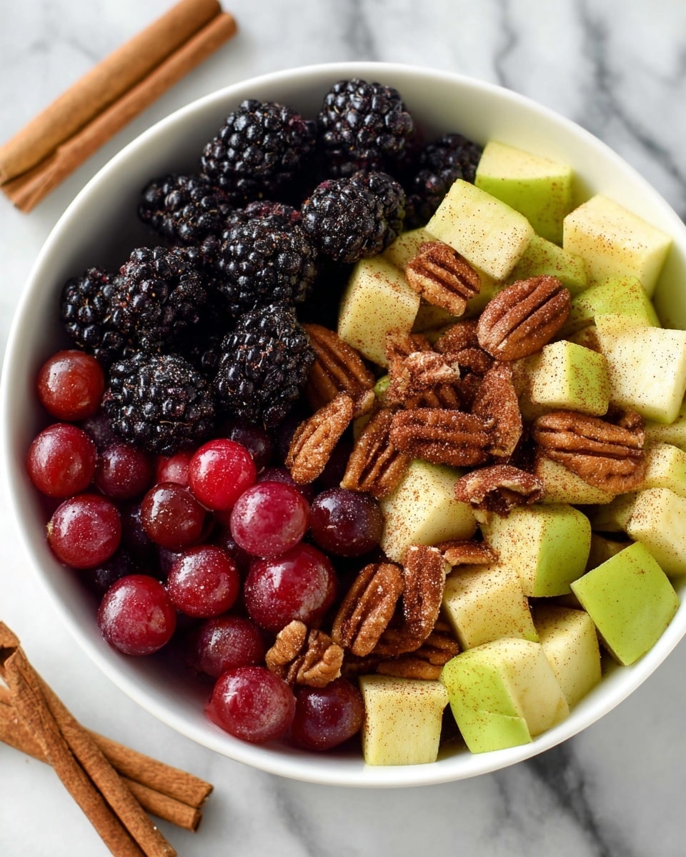 A white bowl filled with three main layers of fresh fruit and nuts carefully arranged side by side. The first layer on the left is shiny, plump dark blackberries with a bumpy texture, next to bright red grapes that look smooth and juicy. The middle layer consists of pale green apple cubes with a dusting of fine brown cinnamon powder, giving a soft textured look. Scattered among and on top are toasted pecans, medium brown with a slightly glossy surface and ridged texture. The bowl is placed on a white marbled surface with two cinnamon sticks lying to the side. Photo taken with an iphone --ar 4:5 --v 7