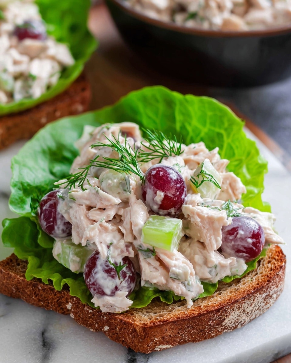 The image shows an open sandwich on a slice of brown bread with a fresh green lettuce leaf on top. Over the lettuce, there is a creamy chicken salad with white and light pink shredded chicken pieces mixed with red grape halves and light green celery chunks. The salad is garnished with small sprigs of fresh green dill. The dish is placed on a white marbled surface, with a blurred bowl of more chicken salad in the background. Photo taken with an iphone --ar 4:5 --v 7