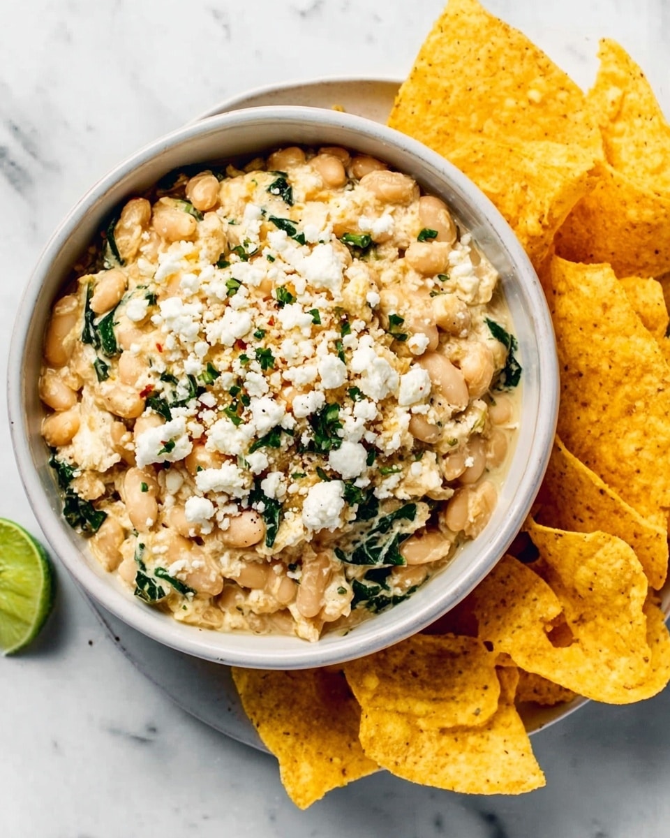 The image shows a white bowl filled with a creamy mixture of white beans and chopped greens, topped with small pieces of crumbled white cheese. The bowl is placed on a white marbled surface, next to a pile of yellow, curved corn chips arranged loosely on the right side of the bowl. A small part of a green lime wedge is visible at the top left corner of the bowl. photo taken with an iphone --ar 4:5 --v 7