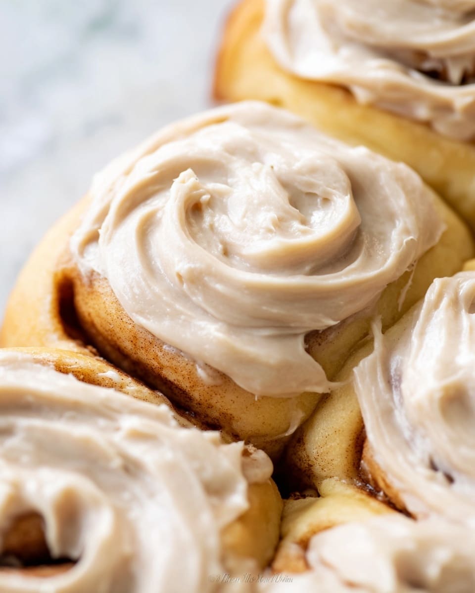 The image shows a close-up of several cinnamon rolls layered side by side, each topped with a thick swirl of creamy beige frosting that looks smooth and slightly textured. The soft dough underneath is light golden yellow, slightly browned in places, with the rolls tightly packed. The frosting covers the top surfaces of each roll but leaves some dough edges exposed, showing a contrast between the creamy topping and fluffy bread. The background is a white marbled texture. Photo taken with an iphone --ar 4:5 --v 7