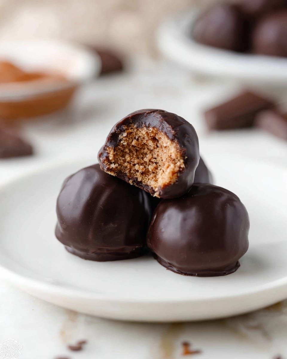 Three dark brown chocolate-covered balls are placed on a white plate with a smooth surface. One ball is on top of two others, and it has a bite taken from it, showing an inner layer of light brown, crumbly, slightly rough texture. The chocolate coating is shiny and smooth but shows slight natural unevenness. The background is softly blurred with a white marbled surface and some blurry chocolate pieces and a small bowl in soft brown and white tones. photo taken with an iphone --ar 4:5 --v 7