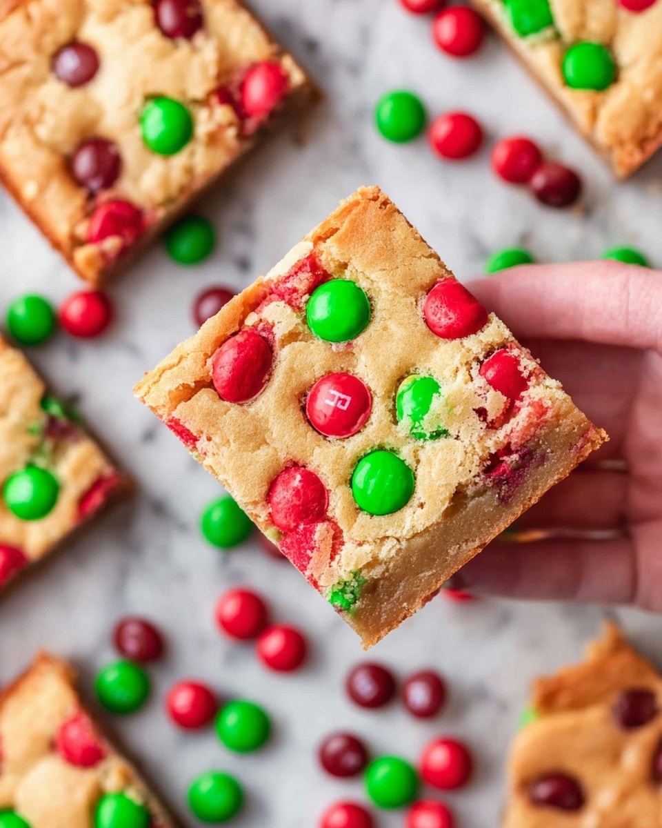A square blondie bar is held by a woman's hand on the right side of the image, showing its top layer filled with red and green candy-coated chocolate pieces embedded on a golden-brown, soft, and slightly cracked surface. The blondie has a thick base with a denser texture visible on the edges. Below the bar, there are scattered red and green candies matching the ones in the blondie, spread casually on a white marbled texture. Several other blondie pieces with the same candy toppings are partially shown in the background, arranged in a loose grid. The image is well-lit with vibrant colors, highlighting the festive look of the dessert. photo taken with an iphone --ar 4:5 --v 7