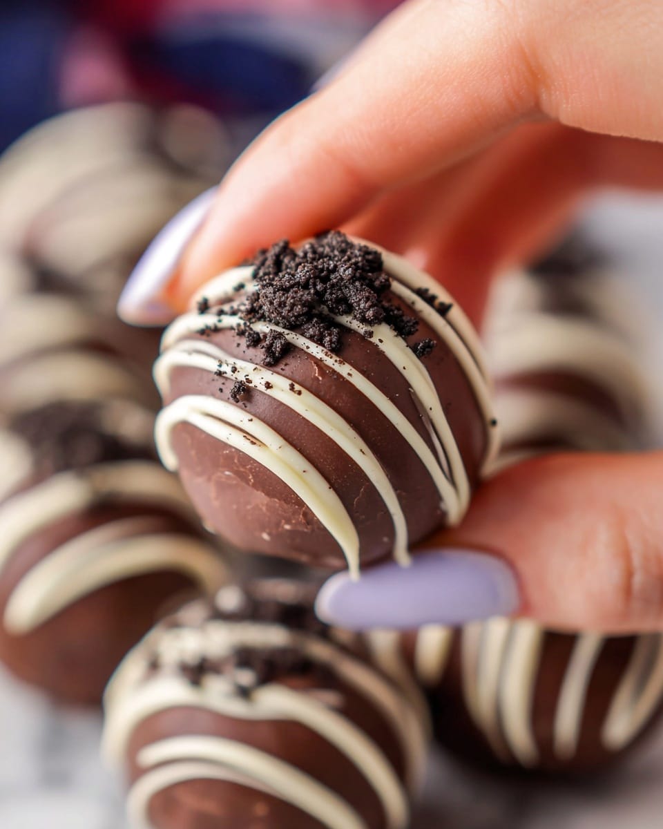 A close-up image of a round chocolate truffle being held gently by a woman's hand with light skin tone and light lavender nail polish. The truffle has a smooth, dark brown chocolate coating with thin white chocolate drizzle forming vertical lines and a sprinkling of fine dark cookie crumbs on top. Several more truffles with similar decoration are blurred in the background, all resting on a white marbled surface. photo taken with an iphone --ar 4:5 --v 7