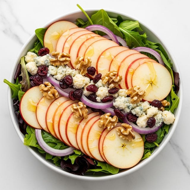 A white bowl filled with a layered fresh salad on a white marbled surface. The base layer is a mix of green leafy lettuce and dark leafy greens. On top are thinly sliced, round apple pieces arranged in small stacks, their creamy white and red edges clearly visible. Scattered between the apple slices are clusters of crumbled blue cheese, small dark red dried cranberries, thin slices of light purple onion, and whole brown walnut halves, adding varied colors and textures. The salad is lightly drizzled with golden honey or dressing, giving a slight shine to the apple slices. Photo taken with an iphone --ar 4:5 --v 7