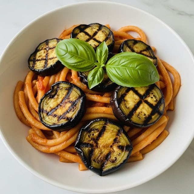 A white bowl filled with short, thick pasta coated in a bright red tomato sauce, mixed with slices of roasted eggplant showing golden-brown char marks and soft texture; two fresh green basil leaves are placed on top, adding a pop of color against the warm tones of the pasta and sauce, all set on a white marbled surface. photo taken with an iphone --ar 4:5 --v 7