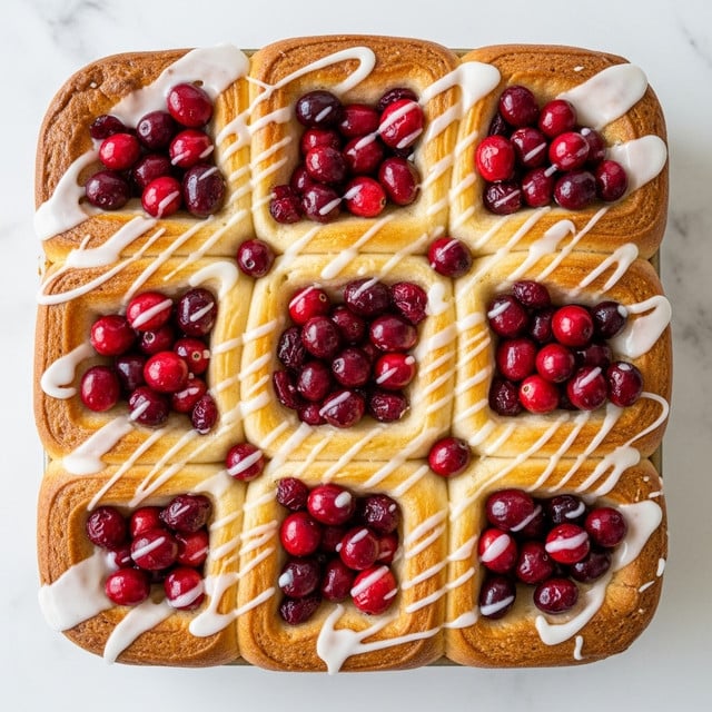 A square pan filled with soft, golden brown pull-apart rolls forming a thick base layer. The rolls are generously topped with a mix of bright red and deep red cranberries, adding vibrant pops of color. A shiny, white glaze is drizzled unevenly over the rolls and berries, creating a glossy finish that contrasts with the warm, fluffy bread texture. The pan sits on a white marbled surface, emphasizing the warm colors of the rolls and the rich reds of the fruit. photo taken with an iphone --ar 4:5 --v 7