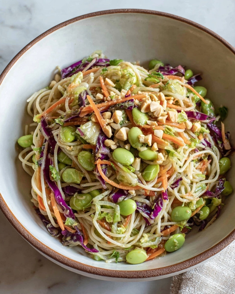 A white bowl with a brown rim filled with a colorful mix of thin white noodles, bright green edamame beans, light green chopped lettuce, thin orange carrot strips, dark purple cabbage slices, chunks of cucumber, and scattered light brown peanuts, all mixed evenly together. The bowl sits on a white marbled surface with a beige cloth and visible chopsticks placed beside it. photo taken with an iphone --ar 4:5 --v 7