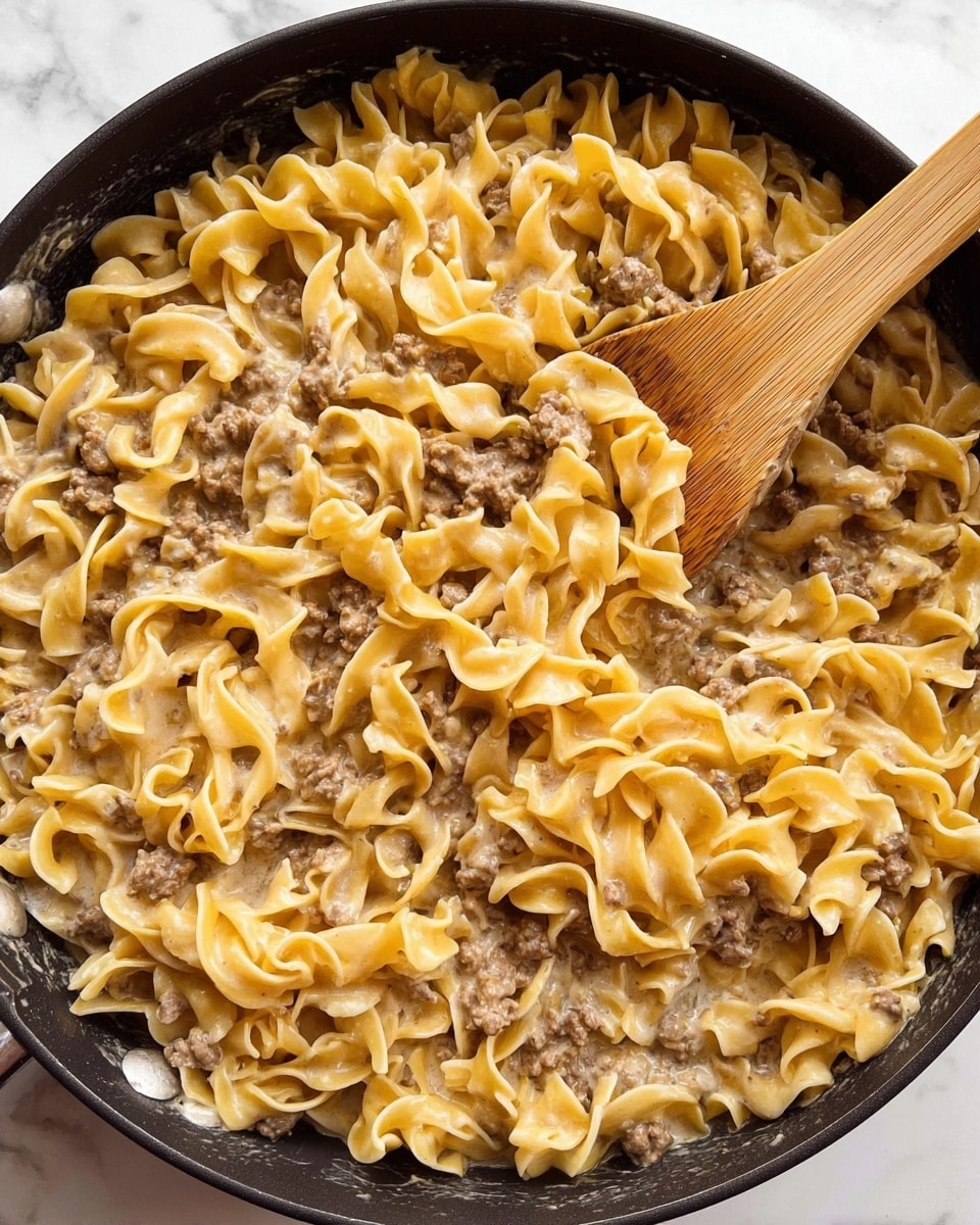 A close-up view of a large black skillet filled with creamy egg noodles mixed with browned ground meat. The noodles are light yellow, soft, and slightly curled with some specks of green herbs scattered throughout. The ground meat pieces are small, browned, and evenly spread among the noodles, all coated in a smooth, light beige creamy sauce. A wooden spoon is partially stirring the mixture from the left side, its textured handle visible. The skillet rests on a white marbled surface. photo taken with an iphone --ar 4:5 --v 7