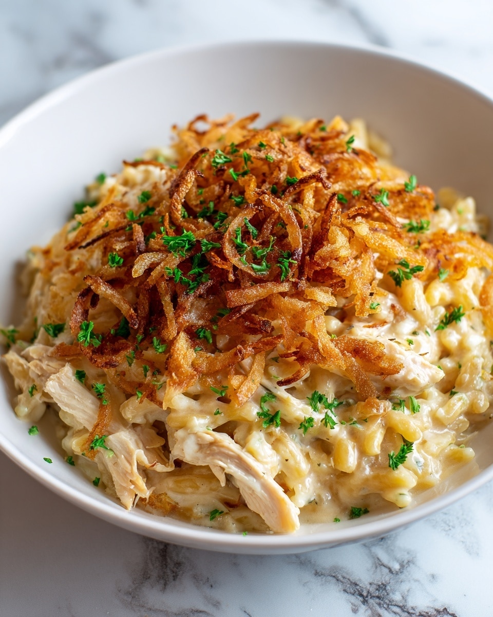 A white bowl filled with a creamy pasta dish that has about three layers: the bottom layer is soft, pale yellow orzo pasta mixed with chunks of light golden chicken; the middle layer is creamy cheese sauce that coats the pasta and chicken, giving a smooth, shiny texture; the top layer is a heap of crispy, golden brown fried onions scattered evenly, adding crunch and contrast, with small bright green parsley pieces sprinkled on top for color. The bowl sits on a white marbled surface, photo taken with an iphone --ar 4:5 --v 7