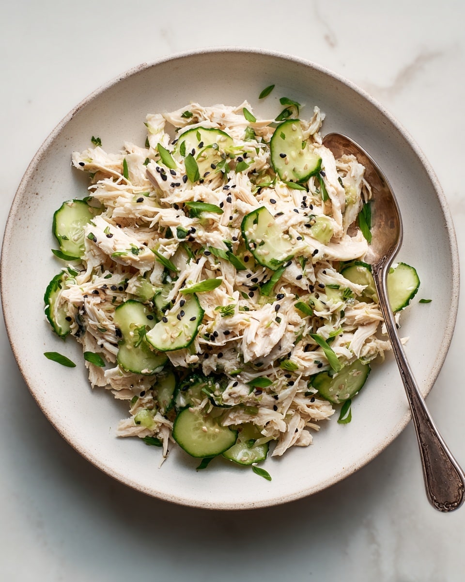 The image shows a white marbled textured background with a white plate filled with a salad made of shredded light beige chicken mixed with chopped green cucumber pieces and sprinkled with small black sesame seeds. Some green herbs are lightly scattered over the dish, adding a fresh touch. A silver spoon is partially buried in the salad on the right side of the plate, with some salad spilling slightly over the edges. The colors create a fresh and simple look, highlighting the contrast between the pale chicken and the vibrant green cucumbers. Photo taken with an iphone --ar 4:5 --v 7