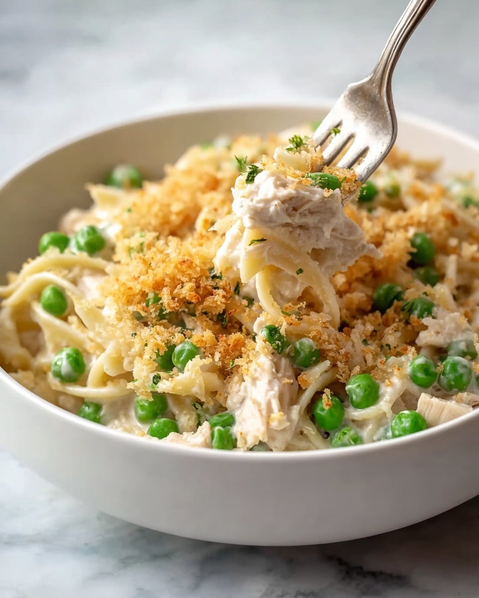 A white bowl filled with creamy pasta layered at the bottom with smooth, pale fettuccine noodles, dotted generously with bright green peas scattered throughout. On top of the pasta is a thick, creamy white sauce mixed with tender chunks of light-colored chicken. The dish is finished with a golden-brown crispy breadcrumb topping that adds texture to the smoothness. A fork with a shiny silver handle lifts a bite showing all the layers together, against a white marbled textured background. photo taken with an iphone --ar 4:5 --v 7
