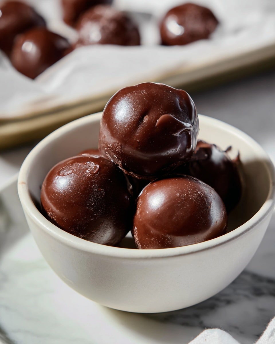 Four dark brown chocolate balls with a smooth, shiny surface sit piled inside a white bowl with a slightly rough texture. Each chocolate ball is round but imperfect, with small ridges and swirls on the coating. The bowl is placed inside another white bowl, both resting on a white marbled surface. In the background, there is a tray lined with parchment paper holding more chocolate balls, slightly out of focus. The lighting creates soft shadows and highlights on the glossy chocolate coating. Photo taken with an iphone --ar 4:5 --v 7