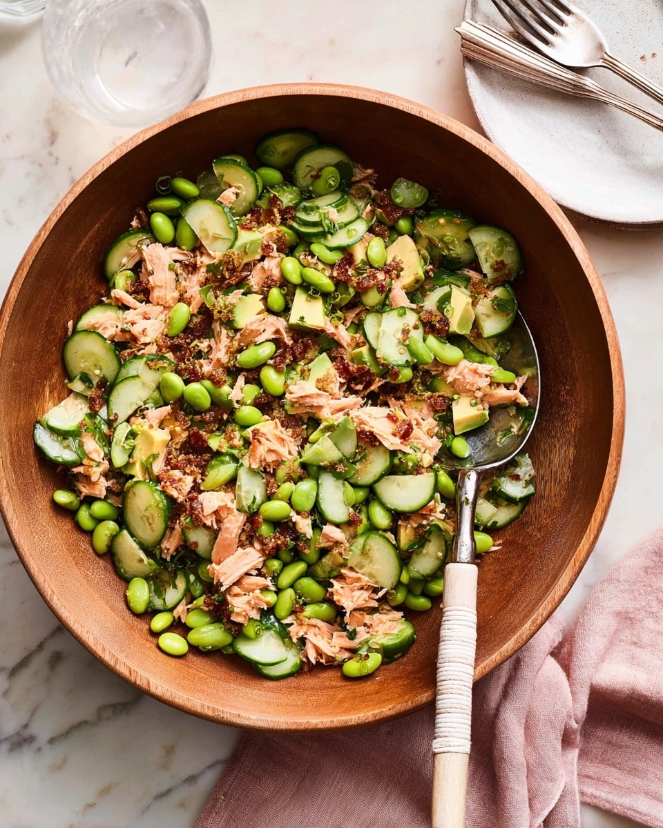 A large wooden bowl filled with a fresh salad showing a mix of thinly sliced green cucumbers, bright green edamame beans, creamy light green avocado chunks, pinkish shredded salmon, and crispy browned bits scattered throughout. A large silver spoon with a wrapped light handle rests inside the bowl. The bowl sits on a white marbled surface, next to a white plate with two silver forks and a pale pink cloth napkin. Photo taken with an iphone --ar 4:5 --v 7