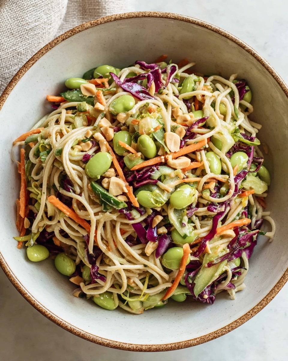 A white bowl with a brown rim filled with a colorful noodle salad sits on a white marbled surface. The salad has several layers, starting with a base of thin, pale beige noodles tangled throughout. Mixed in are bright green edamame beans and small pieces of crunchy chopped lettuce. There are thin strips of orange carrot and purple cabbage scattered evenly above the noodles, adding vivid colors. Small chunks of light green cucumber and smashed peanuts are sprinkled over the top, giving texture and contrast. The salad looks fresh, vibrant, and mixed well to show all ingredients clearly. Photo taken with an iphone --ar 4:5 --v 7