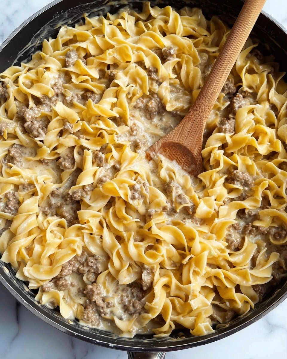 A close-up image of a black pan filled with cooked egg noodles mixed with ground beef in a creamy sauce. The noodles are yellowish with a soft, slightly wavy texture, and the ground beef pieces are small and brown, evenly spread throughout. The creamy sauce lightens some parts of the dish with a smooth, glossy appearance. A wooden spoon is placed inside the pan, partially covered with the noodles and sauce. The background shows a white marbled surface. photo taken with an iphone --ar 4:5 --v 7
