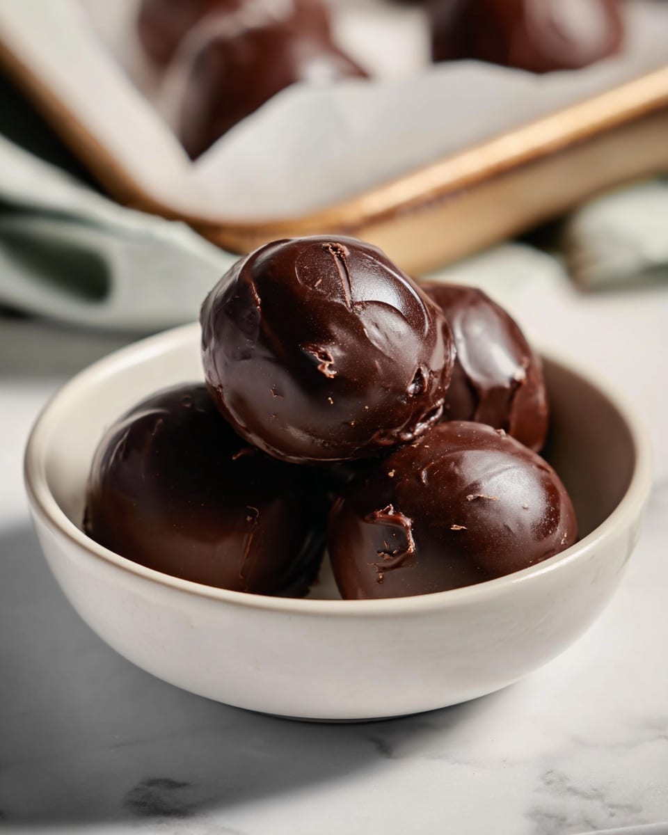 The image shows four smooth, shiny chocolate balls with a glossy dark brown coating, each slightly irregular in shape and nestled closely together in a white bowl. The chocolate surface is rich with subtle textures and soft reflections of light. The bowl has a simple, smooth finish and sits inside another white bowl, both resting on a white marbled surface. In the background, more chocolate balls can be seen in a tray lined with parchment paper, blurred softly to keep the focus on the bowl in the foreground. photo taken with an iphone --ar 4:5 --v 7