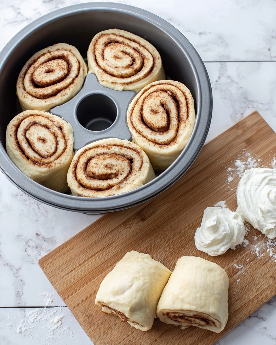 The image shows six raw cinnamon rolls with a swirl pattern of light beige dough and darker brown cinnamon filling. Four rolls are neatly placed inside a dark gray fluted bundt pan at the bottom right of the image, showing the spiral layers clearly from above. Beside the pan, on a wooden cutting board on a white marbled surface, two more cinnamon rolls are stacked, and two dollops of white cream cheese frosting sit nearby in soft, rounded shapes. The dough looks soft and slightly fluffy, while the cinnamon filling has a fine textured look. Photo taken with an iphone --ar 4:5 --v 7