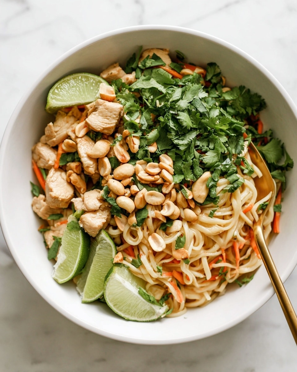 A bowl filled with three main visible layers: the bottom layer is light beige cooked noodles, the middle layer consists of small pieces of cooked chicken and shredded orange carrots mixed evenly, and the top layer is a mix of green fresh cilantro leaves, light brown whole peanuts scattered around, and two lime wedges placed to the side inside the bowl; a gold fork is inserted into the noodles on the right side of the bowl. The bowl is white and placed on a white marbled surface. Photo taken with an iphone --ar 4:5 --v 7