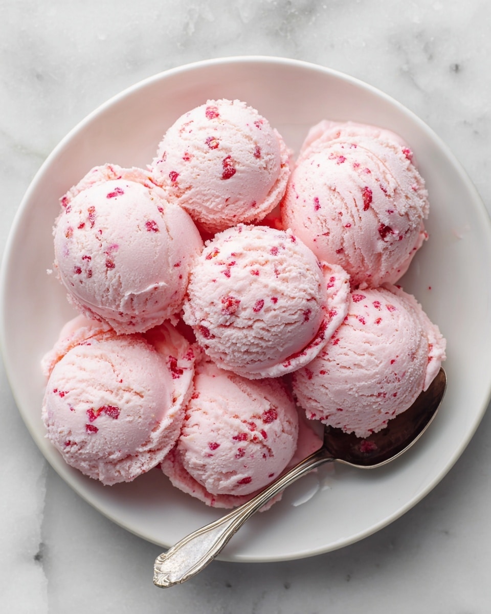 A white bowl filled with eight rounded scoops of light pink ice cream with small darker pink bits mixed inside, giving it a textured look. The ice cream appears creamy and smooth with slight cracks on the surface of each scoop. A silver spoon rests inside the bowl on the right side, partially under the ice cream. The bowl sits on a white marbled surface, adding a clean and bright background to the image. photo taken with an iphone --ar 4:5 --v 7