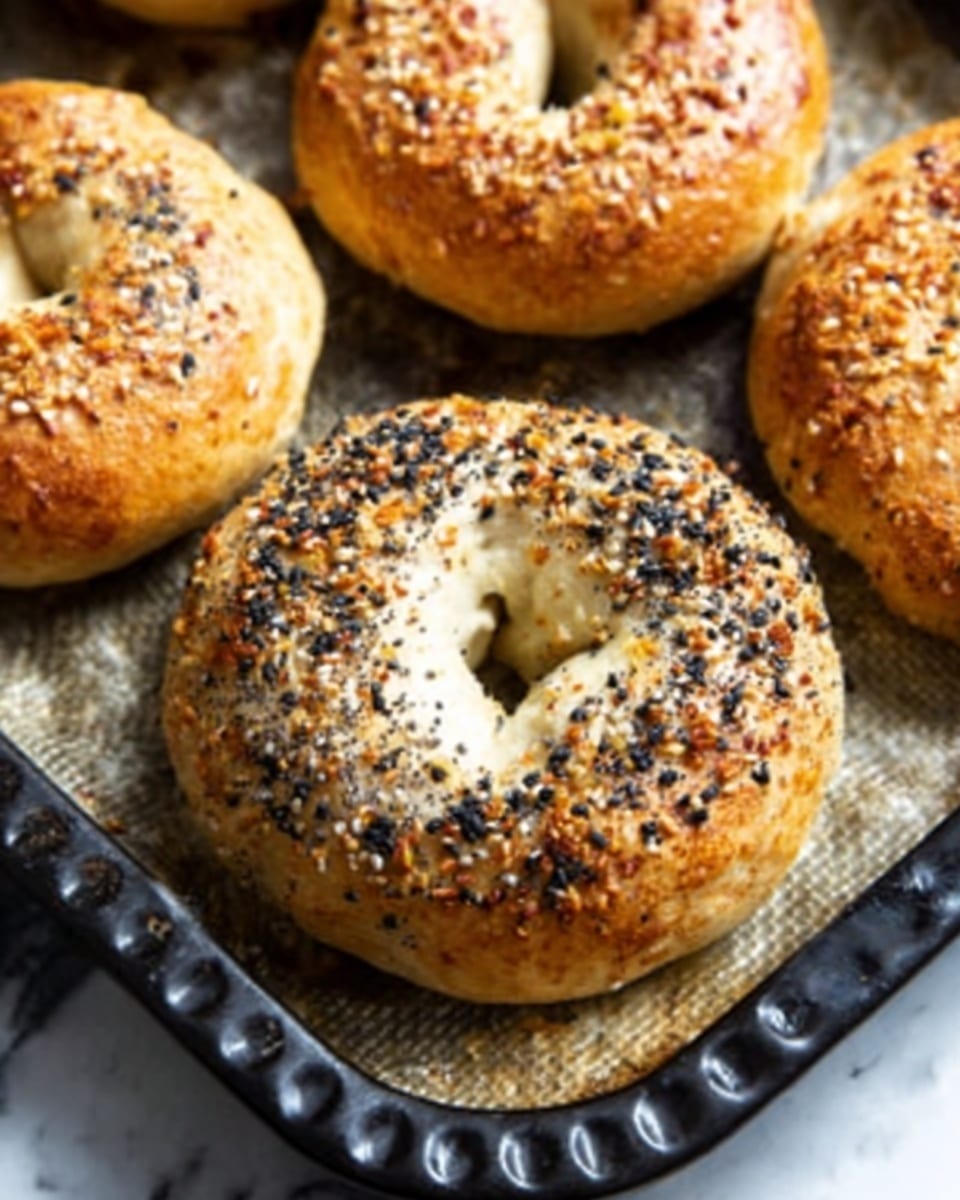 The image shows a close-up of four golden-brown bagels on a baking tray with a dark surface. The main bagel in focus is sprinkled with mixed seeds like sesame, poppy, and salt, giving it a speckled look with white, black, and light brown colors on the top crust. The bagel crust looks shiny and smooth, with a slightly rough texture from the seeds. The other bagels in the background have a plain golden crust with a few shiny spots. The baking tray has a slightly worn, dark metal texture. photo taken with an iphone --ar 4:5 --v 7