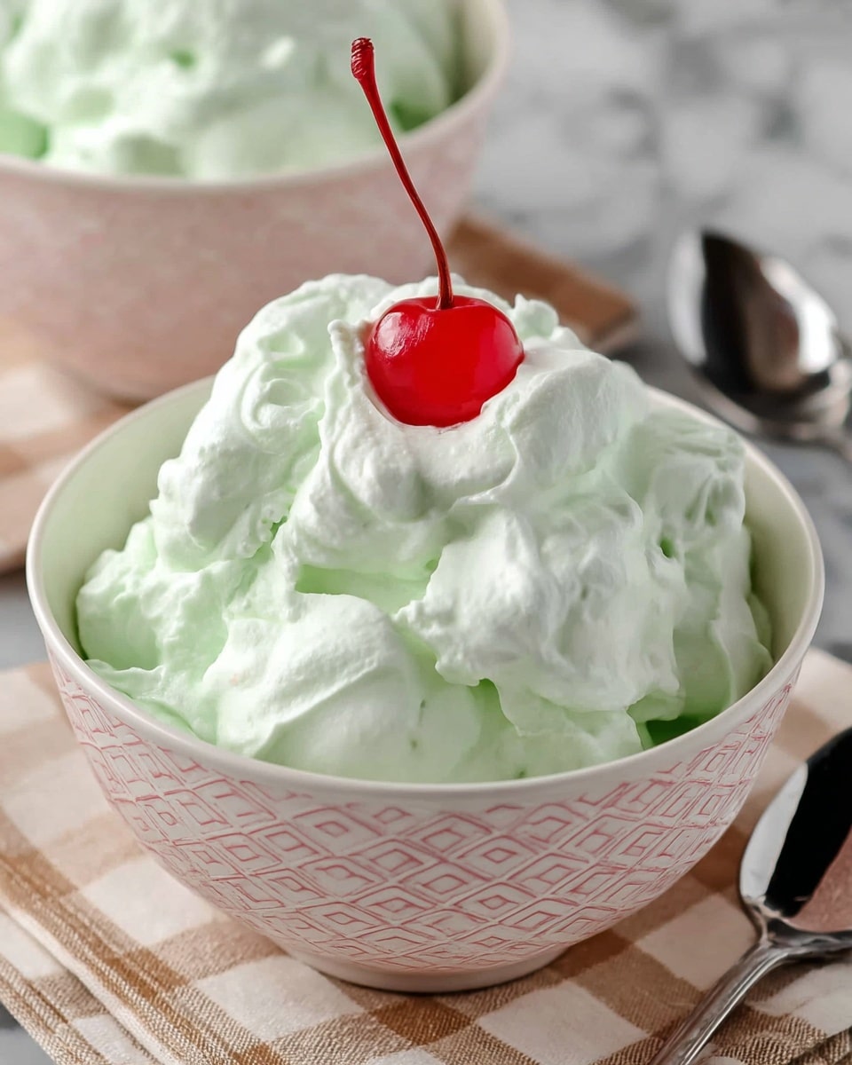 The image shows a close-up of a round white bowl with a light pink geometric pattern, filled with a fluffy light green whipped dessert that has a soft, smooth texture with some peaks. On top of the whipped dessert, there is a single bright red cherry with a long stem adding contrast to the pale green. The bowl sits on a beige and white checkered cloth on a white marbled surface, with a second similar bowl blurred in the background. A silver spoon is partially visible to the right of the bowl. Photo taken with an iphone --ar 4:5 --v 7