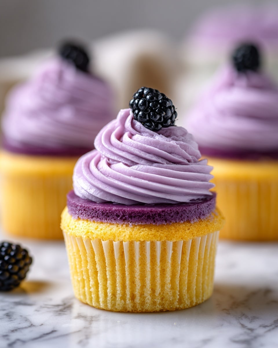 The image shows a close-up of a purple cupcake with three main layers. The bottom layer is a golden yellow cake wrapped in a light purple cupcake liner. On top of the cake, there is a smooth, glossy dark purple jelly layer. The top layer is a swirl of soft, light purple frosting with a creamy texture, and it is decorated with a single shiny black blackberry. In the background, two more identical cupcakes are slightly out of focus, sitting on a white marbled surface. photo taken with an iphone --ar 4:5 --v 7