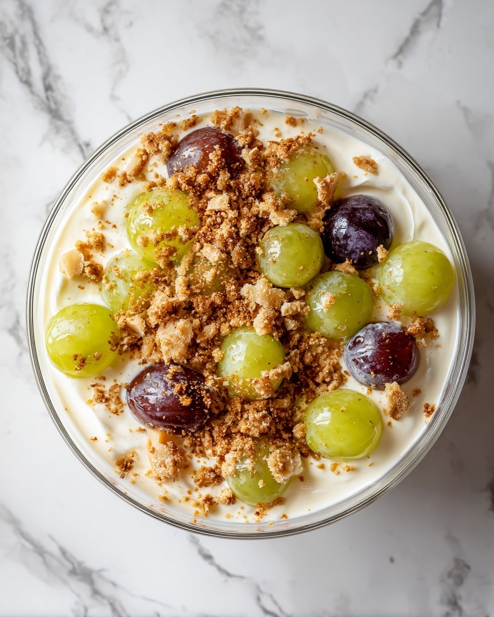This image shows a clear glass bowl filled with a creamy dish made of two main layers: the first layer is white, smooth cream that coats the second layer of whole grapes, both green and purple. On top, there is a generous sprinkle of crushed nuts and light brown crumbs which add a rough texture and cover the cream and grapes partially. The bowl is placed on a white marbled surface, and the grapes peek through the glossy cream, giving a fresh and rich look. photo taken with an iphone --ar 4:5 --v 7
