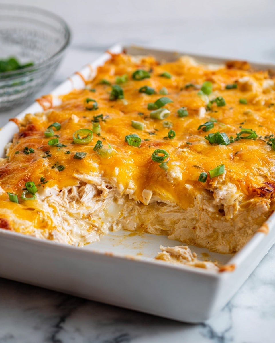 A close-up view of a white rectangular baking dish filled with a creamy layered casserole. The top layer is melted golden orange cheese with small, finely chopped green onions sprinkled evenly across the surface. A portion has been scooped out, showing an inner layer of shredded chicken mixed with a smooth, thick white sauce or cream. The casserole looks soft and cheesy, with the chicken layer beneath the bright cheese top. The dish is set on a white marbled surface, and a woman's hand is holding a large spoon scooping from the dish. Photo taken with an iphone --ar 4:5 --v 7