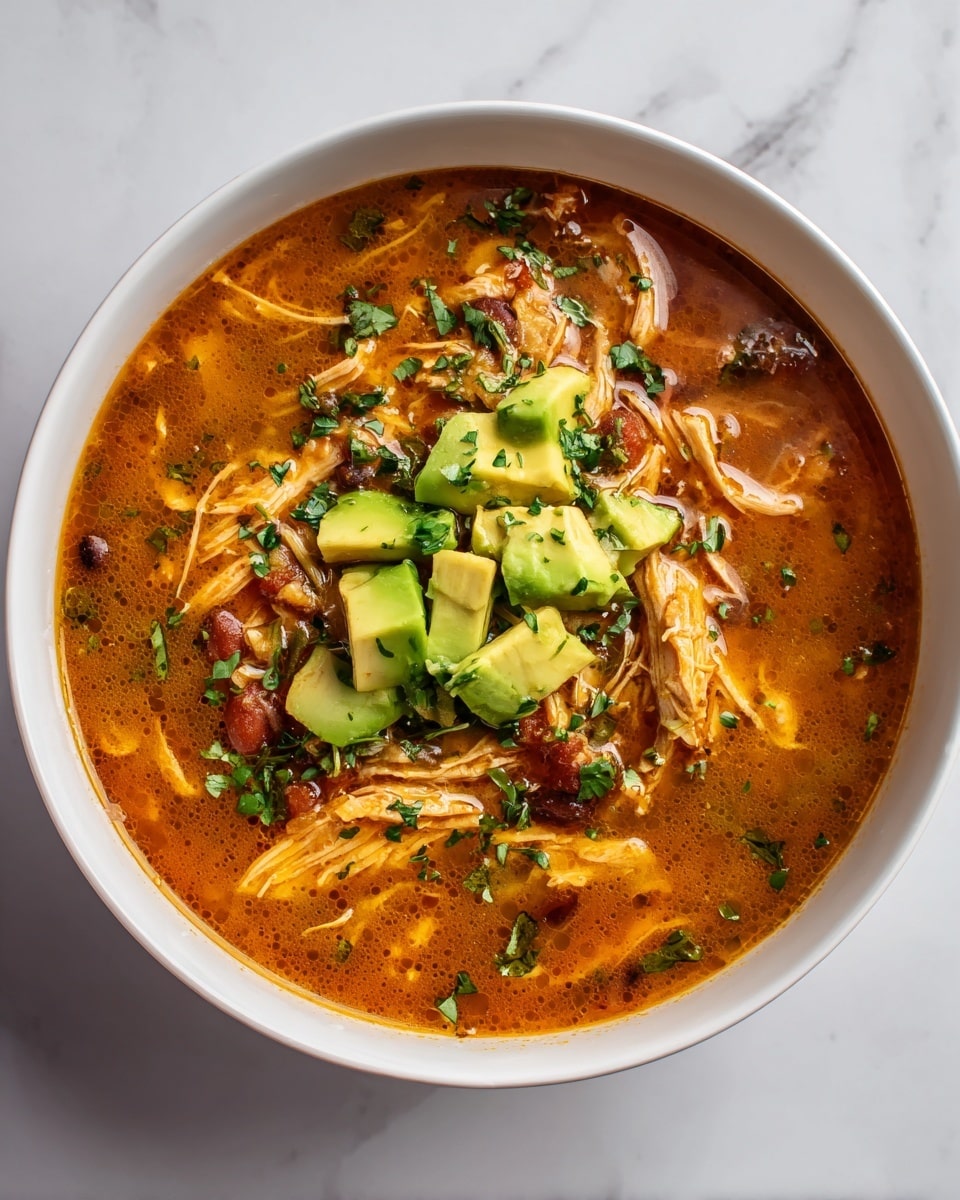 A close-up image of a thick stew inside a white pot with a green rim, showing multiple layers of ingredients. The bottom layer is a rich reddish-orange broth with visible bits of tomato and spices. On top, there are shredded pieces of chicken spread throughout, mixed with black beans scattered evenly. Small cubes of bright green avocado sit on the surface, along with freshly chopped cilantro leaves sprinkled generously, adding a fresh green touch. The textures range from smooth broth to chunky beans and soft chicken, creating a hearty and colorful dish. The background is a white marbled texture. Photo taken with an iphone --ar 4:5 --v 7