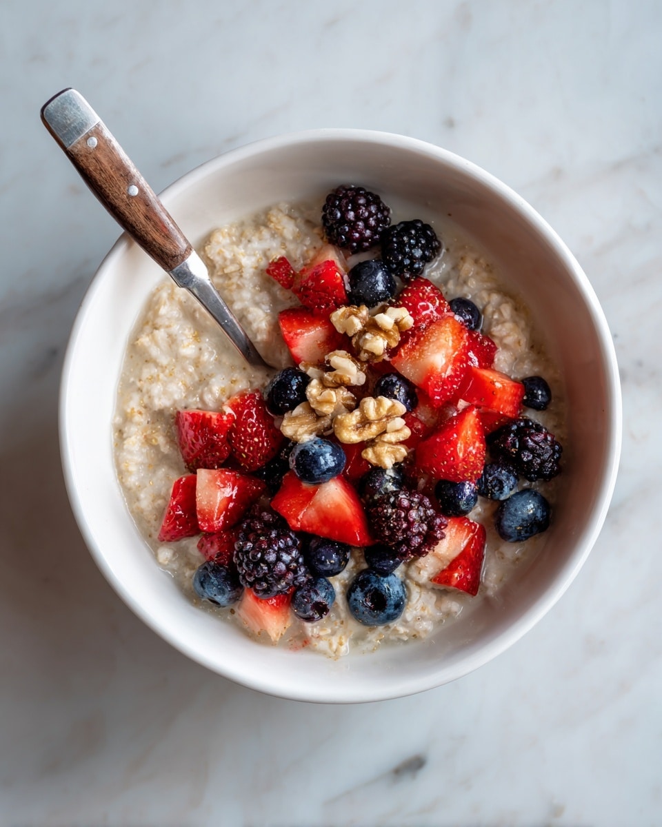 A white bowl filled with creamy oatmeal as the base layer, light beige in color with a soft, slightly lumpy texture. The oatmeal is topped with a colorful mix of fresh berries, including deep black blackberries, dark blue blueberries, and bright red diced strawberries scattered mostly on one side. Small pieces of golden-brown walnuts are sprinkled over the berries, adding a crunchy texture. A silver spoon with a wooden handle rests partially inside the bowl on the right side. The bowl is set on a white marbled surface. photo taken with an iphone --ar 4:5 --v 7