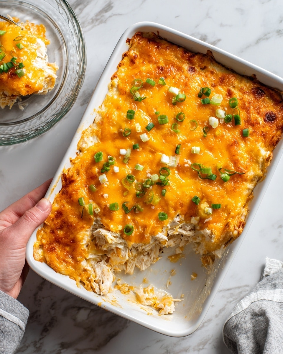 The image shows a baked casserole in a white rectangular dish with layers visible where a piece has been taken out. The bottom layer is creamy with shredded chicken mixed in, topped with melted orange cheddar cheese that covers the surface evenly. Small pieces of green chopped onions are scattered on top of the cheese, adding color contrast. The dish sits on a white marbled surface, and a woman's hand holding a clear glass dish is partially visible. Photo taken with an iphone --ar 4:5 --v 7