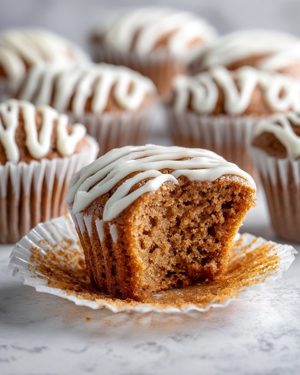 A close-up view of a moist brown muffin with a soft, airy texture and a bite taken out of its top front, revealing its crumbly inside. The muffin is in a white paper liner, and its top is drizzled with creamy white icing that appears smooth and slightly shiny. In the background, there are several similar muffins, each topped with white icing in a zigzag pattern, placed on a white marbled surface. The lighting brightens the muffins’ warm brown color and highlights the icing details. photo taken with an iphone --ar 4:5 --v 7