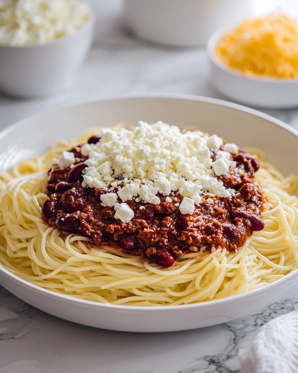 A white plate holds a serving of cooked spaghetti noodles forming the base layer, topped by a thick layer of dark red kidney beans mixed with brown minced meat sauce. On top, there are small white cheese cubes sprinkled generously, adding contrast. The scene is set on a white marbled surface, with a blurred bowl of grated cheese and a small white container in the background. Photo taken with an iphone --ar 4:5 --v 7