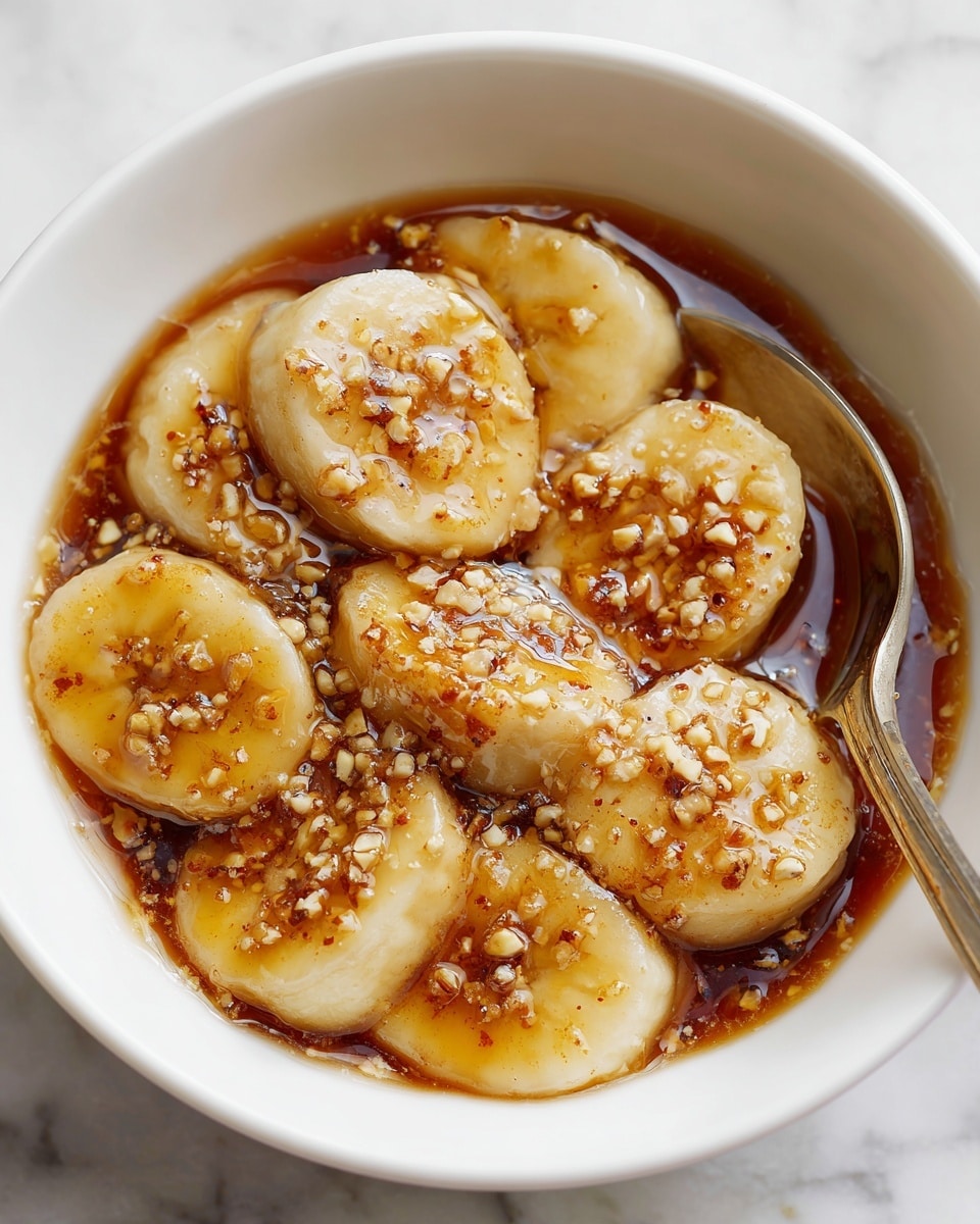 A close-up of a white bowl filled with several thick slices of banana layered flat, covered in a thick, glossy brown syrup, with small bits of crushed nuts sprinkled on top. A silver spoon is resting on the edge of the bowl, partially visible. The background shows a white marbled surface. photo taken with an iphone --ar 4:5 --v 7