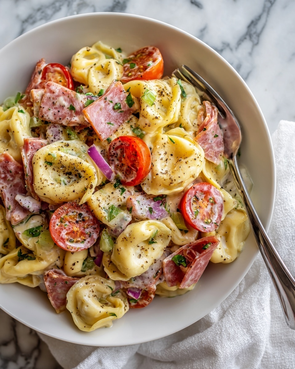 A close-up of a white plate filled with a layered tortellini pasta salad. The first layer shows creamy pale yellow tortellini noodles with a smooth texture. Mixed through are bright red cherry tomato halves, pieces of pale pink ham, thin slices of light green celery, and small white chunks of onion. The dish is lightly sprinkled with coarse ground black pepper. The plate sits on a white marbled surface with a fork resting to the right, and a few blurred greens and a cherry tomato in the background. photo taken with an iphone --ar 4:5 --v 7