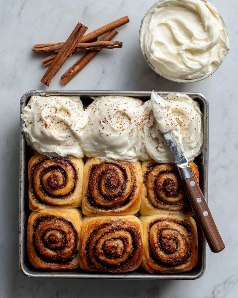 A metal baking pan holds nine pumpkin cinnamon rolls arranged in three rows of three. Each roll has two layers: a bright orange baked dough base with a dark brown cinnamon swirl on top. Five of the rolls are thickly covered with white cream cheese frosting, spread unevenly to show soft texture, and sprinkled with a light dusting of brown cinnamon powder. One roll on the right side has frosting being spread over it by a knife with a wooden handle resting on it. The pan sits on a white marbled surface with cinnamon sticks, a small bowl of white frosting with a swirl, and another small bowl with brown cinnamon powder scattered nearby. Photo taken with an iphone --ar 4:5 --v 7