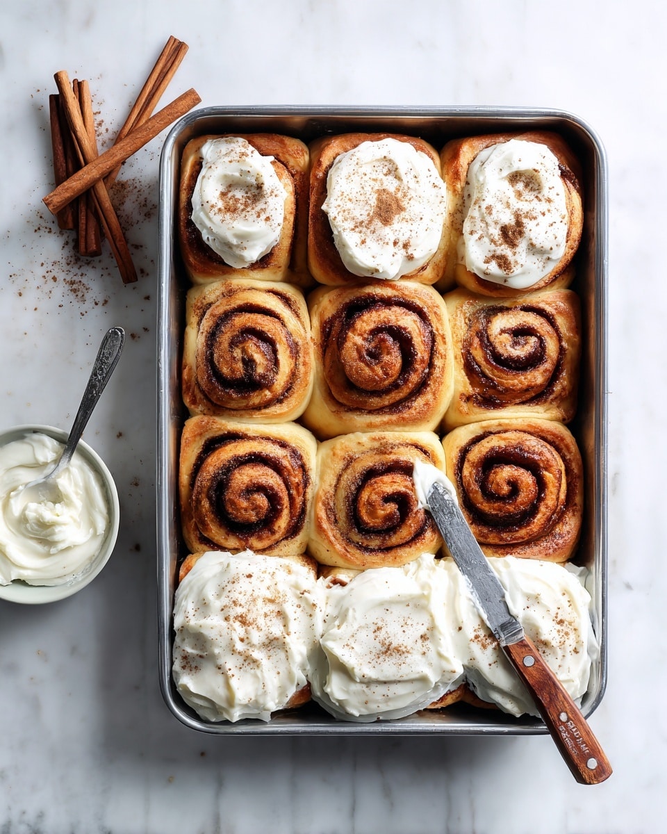 A metal baking tin holds nine golden-brown cinnamon rolls arranged in a 3x3 grid. Each roll shows a dark brown cinnamon swirl pattern on top, with the lower six rolls covered with a thick layer of creamy white frosting sprinkled lightly with cinnamon powder. The three rolls at the top left are partially frosted. A shiny metal knife with a wooden handle rests over the top right roll, spreading a dollop of frosting. To the left of the tin, three cinnamon sticks are placed beside a bowl of smooth white frosting, all set on a white marbled surface. Photo taken with an iphone --ar 4:5 --v 7