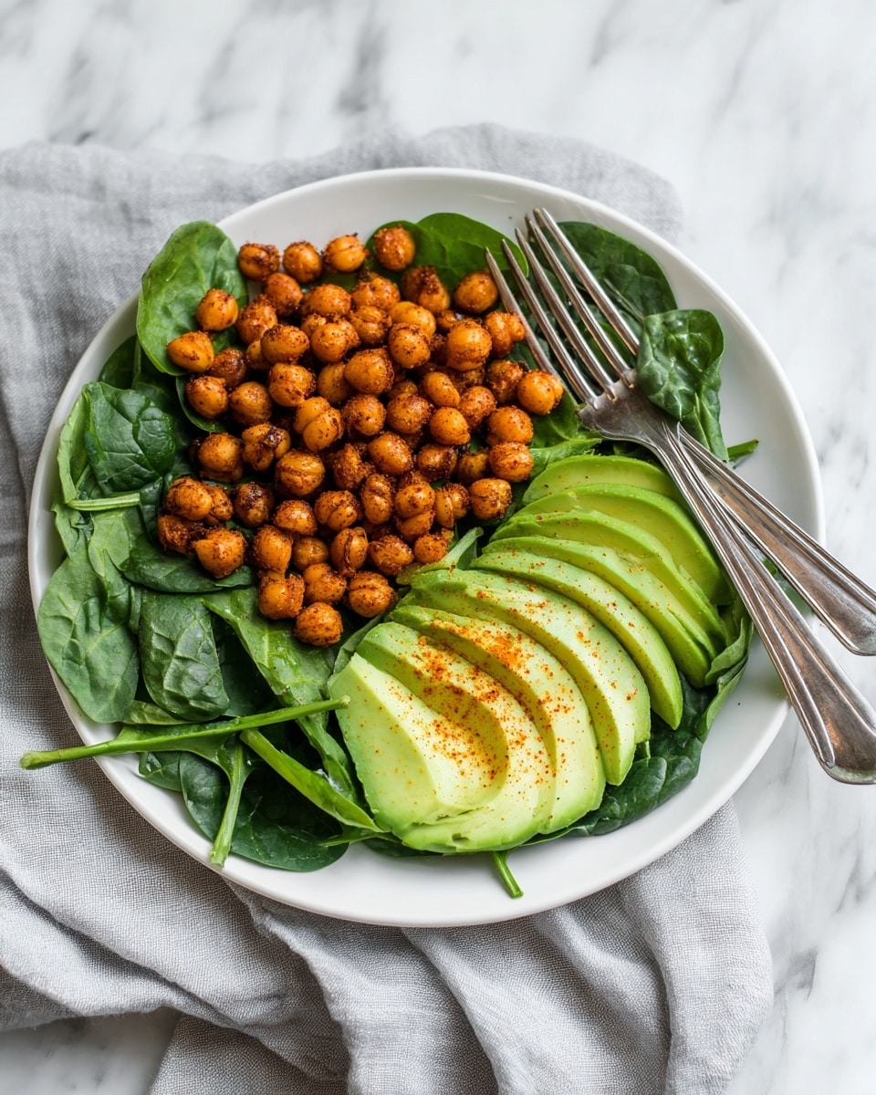 The dish is presented in a white plate with a slightly raised edge, sitting on a white marbled surface with a light cloth nearby. The base layer consists of fresh, glossy spinach leaves that cover the plate and form a vibrant dark green bed. On top of the spinach, there is a generous heap of golden chickpeas, which look soft and slightly oily with a light sprinkle of seasoning that adds a warm color contrast. Arranged neatly on one side of the chickpeas, there are thin, bright green avocado slices that curve gently, adding a smooth texture and fresh look. A silver fork rests beside the food on the edge of the plate, enhancing the simple yet fresh presentation. photo taken with an iphone --ar 4:5 --v 7