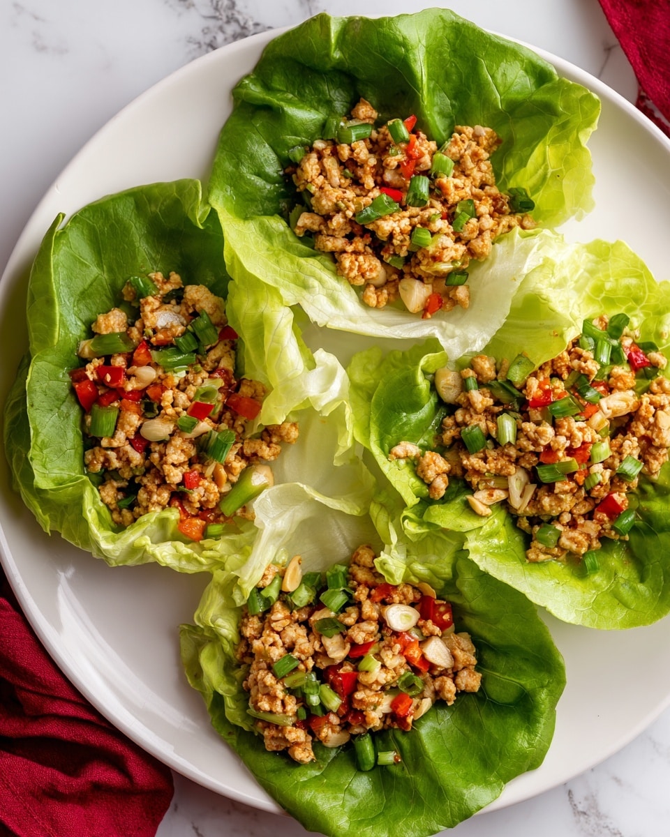 A white plate holds five lettuce wraps arranged in a loose circle, each made of bright green, crisp-looking large lettuce leaves forming a bowl shape. Inside each leaf is a filling of finely chopped, cooked ground chicken mixed with diced red bell peppers and finely chopped green onions, giving a texture of small bits and a mix of light brown, red, and green colors. The plate rests on a white marbled surface with a blurred red cloth nearby. Photo taken with an iphone --ar 4:5 --v 7