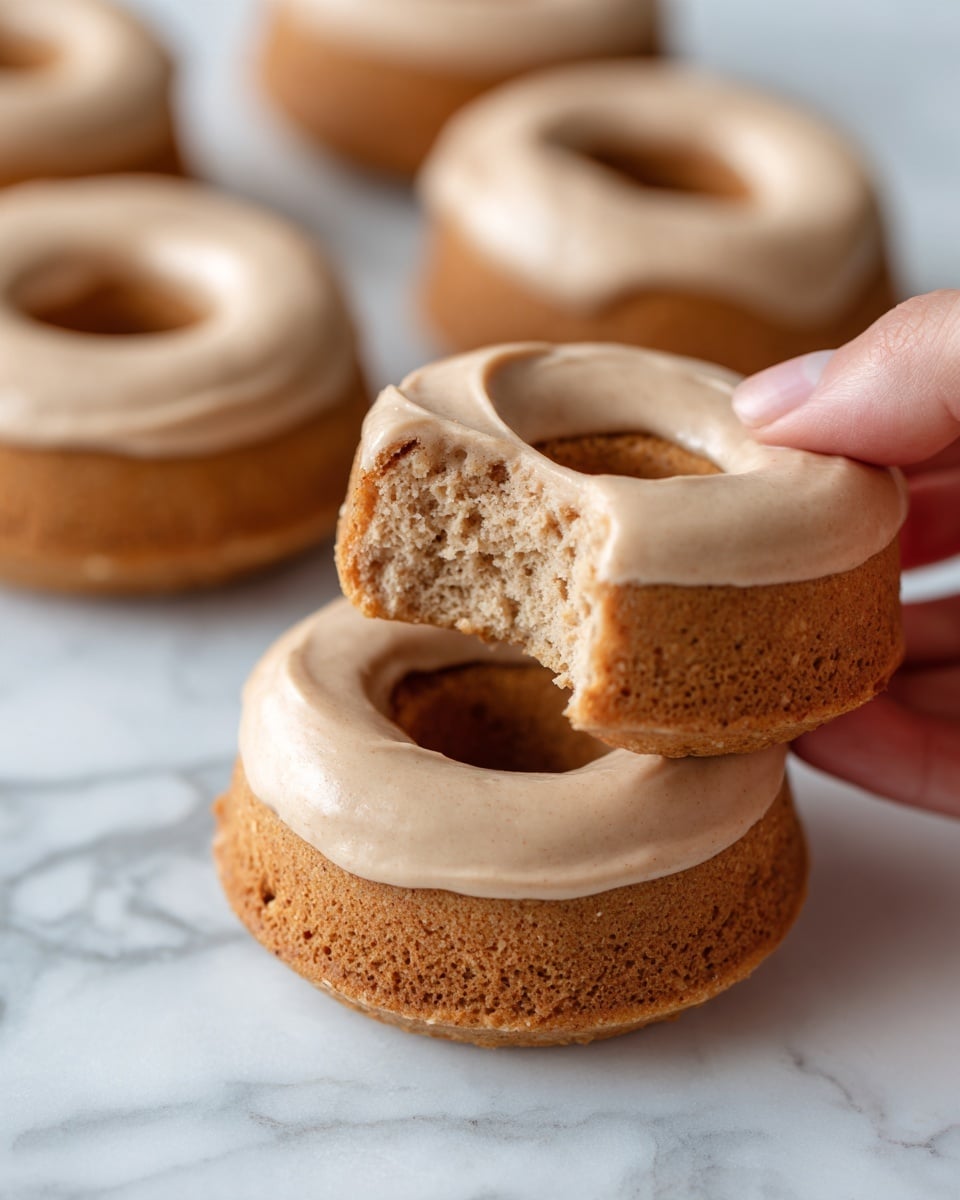 The image shows a single pumpkin donut with light brown color and a soft, crumbly texture. It is topped with smooth, creamy brown sugar frosting that covers the top half, dripping slightly over the edges. A bite is taken out of the donut, revealing the moist inside with tiny air pockets. The donut is placed on a clean white plate on a white marbled surface. The lighting is soft, highlighting the frosting's glossiness and the donut's texture. photo taken with an iphone --ar 4:5 --v 7
